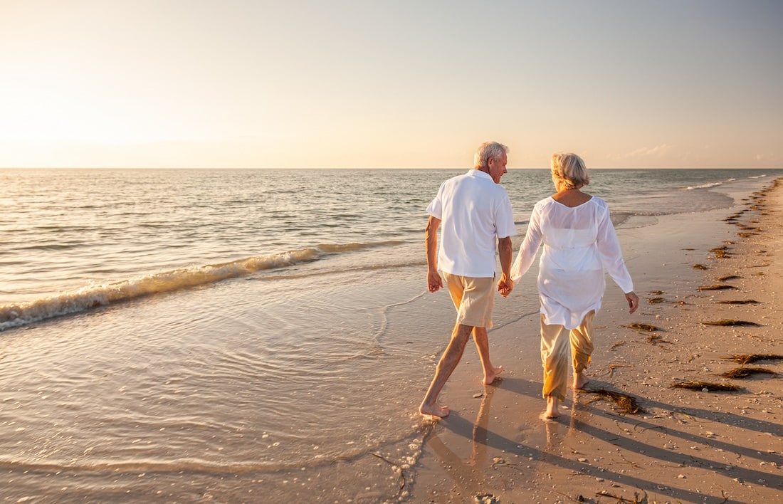 older white couple wearing white summer clothing walking along the beach at sunset.
