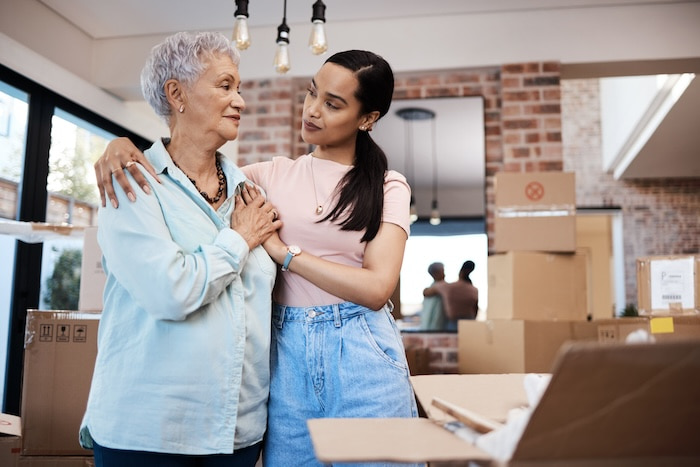 Younger woman comforting her elderly mother with boxes around their home that has moving boxes.
