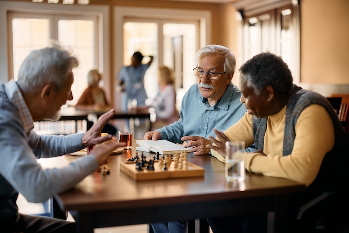 three elderly men sitting around a table playing a mini chess game