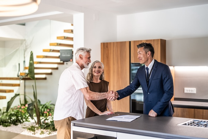 Asian realtor shaking hands with an older man with his wife inside of a new home.