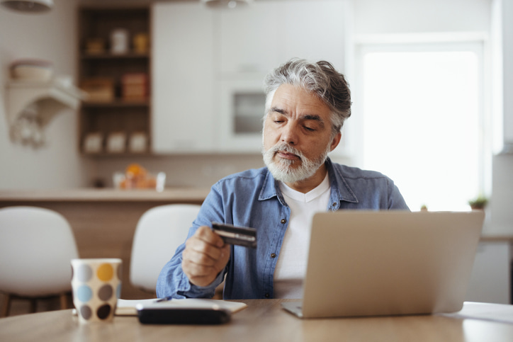 senior man looking at his credit card while at a table with his laptop