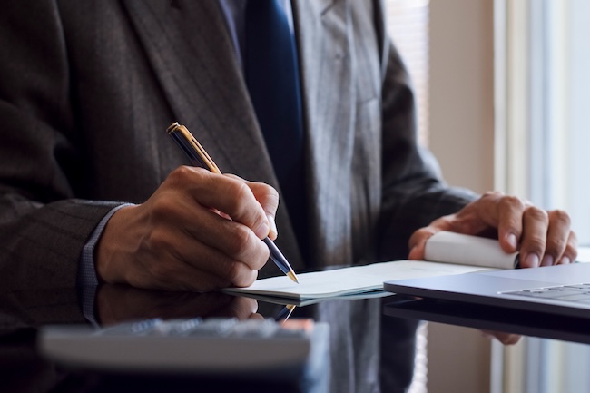 A businessman in a suit is handwriting and signing a chequebook with a calculator on the dark mirror table at the office.