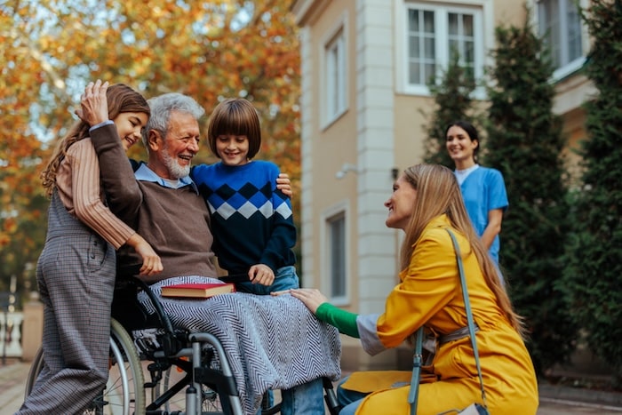 woman kneeling in front of elderly family member in a wheelchair surrounded by younger grandkids
